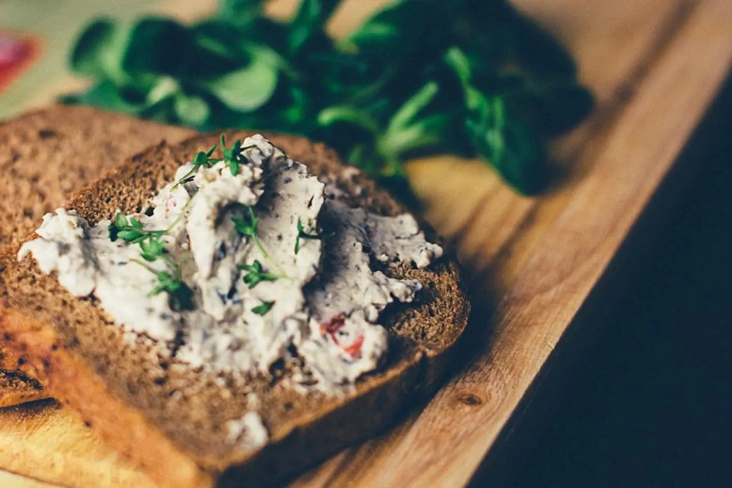 Dunkles Brot mit heller Käsecreme und Kräutern auf einem Holzbrett